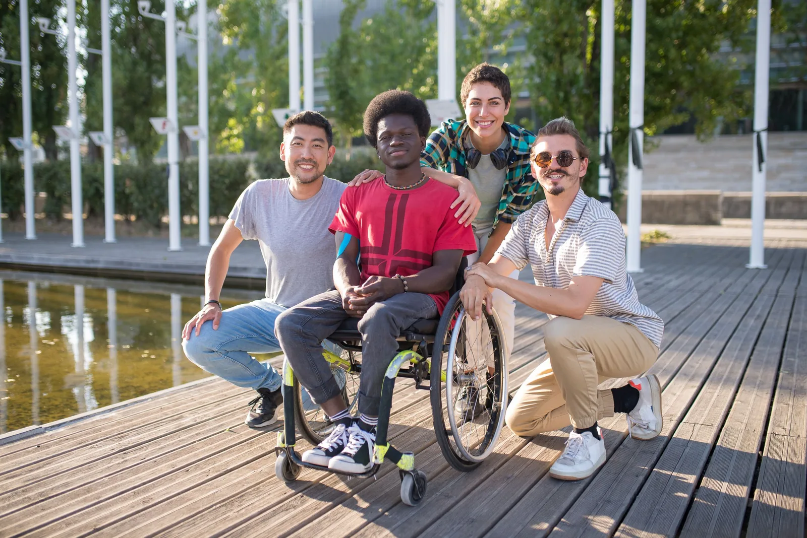Four friends gathered around a man using a wheelchair beside a pond.