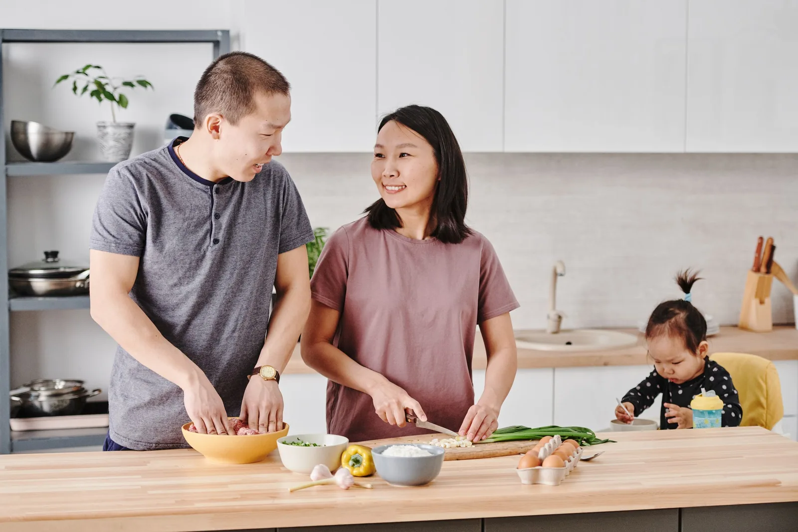 Two adults preparing food together in a kitchen while a child sits nearby.