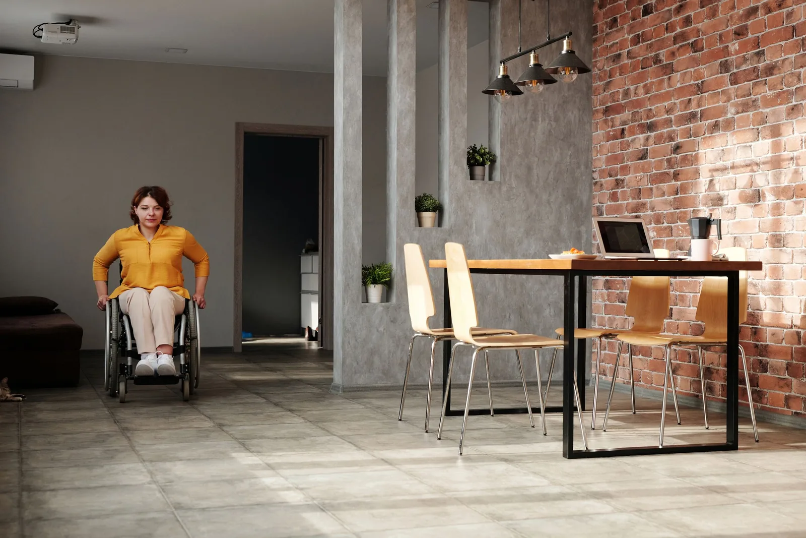 A woman using a wheelchair in a bright room with a dining table and brick wall.